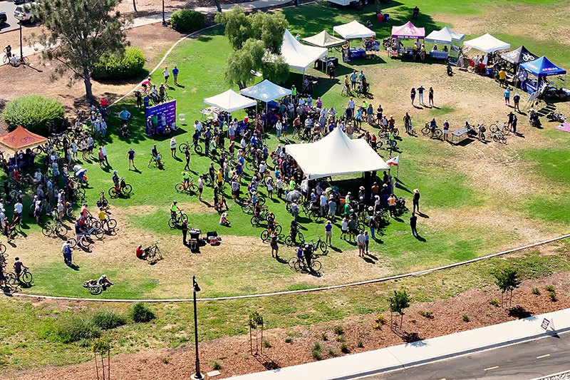 An aerial shot of bike riders and supports gathering around a semicircle of tents on the lawn at Bird Park at Morley Field to celebrate the opening of the Pershing Bikeway and learn more about SANDAG's bikeway and walkway projects.