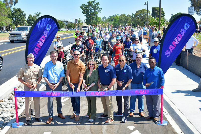 A crowd of people on bikes celebrates behind a small group of officials preparing to cut the ceremonial ribbon. Ribbon cutters include SANDAG Vice Chair and City of San Diego City Council President Sean Elo-Rivera, Executive Director of the San Diego County Bicycle Coalition Chloé Lauer, City of San Diego Mayor Todd Gloria, City of La Mesa Mayor and SANDAG Councilmember Jack Shu, SANDAG CEO Mario Orso, and others. 