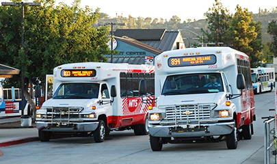 Two red and white MTS minibuses at a transit center during dusk. The number 894 Morena Village minibus is driving by the parked number 833 Santee Trolley minibus.